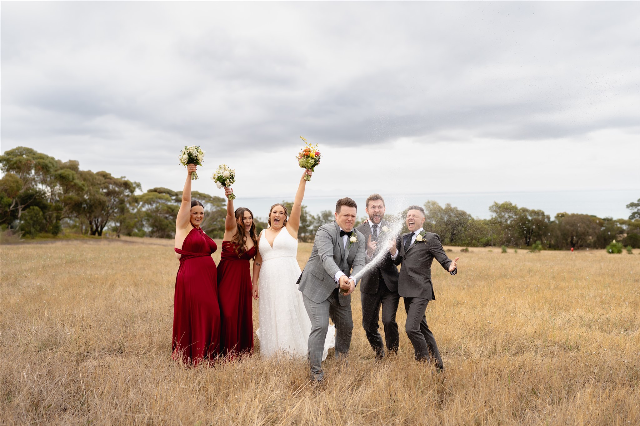Wedding party cheering and walking through the open field at Terindah Estate, with the bride and groom leading the way under a cloudy sky.